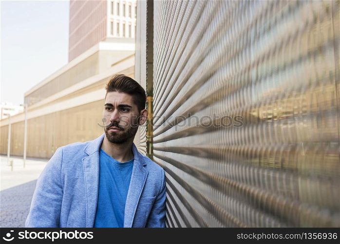 Young bearded man in urban background wearing casual clothes while leaning on a wall and looking aside