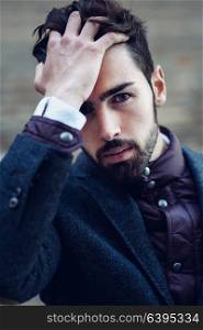 Young bearded man, model of fashion, in urban background wearing british elegant suit. Guy with beard and modern hairstyle touching his hair