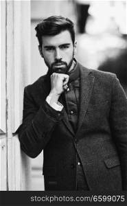 Young bearded man, model of fashion, in urban background wearing british elegant suit. Guy with beard and modern hairstyle in the street.