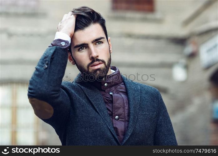 Young bearded man, model of fashion, in urban background wearing british elegant suit. Guy with beard and modern hairstyle touching his hair. Young bearded man wearing british elegant suit touching his hair