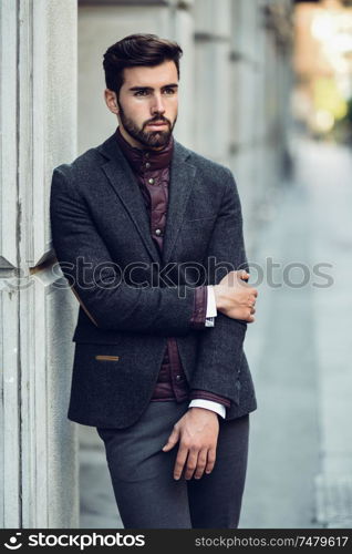 Young bearded man, model of fashion, in urban background wearing british elegant suit. Guy with beard and modern hairstyle in the street.. Young bearded man in urban background wearing british elegant suit in the street.