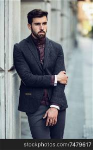 Young bearded man, model of fashion, in urban background wearing british elegant suit. Guy with beard and modern hairstyle in the street.. Young bearded man in urban background wearing british elegant suit in the street.