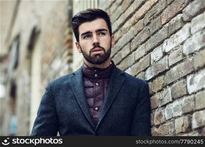 Young bearded man, model of fashion, in urban background wearing british elegant suit. Guy with beard and modern hairstyle in the street.