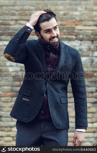 Young bearded man, model of fashion, smiling in urban background wearing british elegant suit. Guy with beard and modern hairstyle in the street.