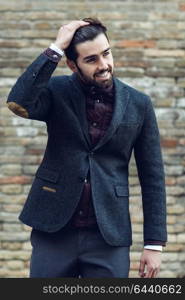 Young bearded man, model of fashion, smiling in urban background wearing british elegant suit. Guy with beard and modern hairstyle in the street.