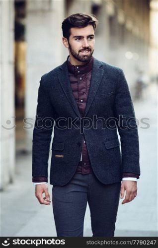 Young bearded man, model of fashion, smiling in urban background wearing british elegant suit. Guy with beard and modern hairstyle in the street.