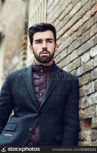 Young bearded man, model of fashion, standing in urban background wearing british elegant suit. Guy with beard and modern hairstyle in the street.