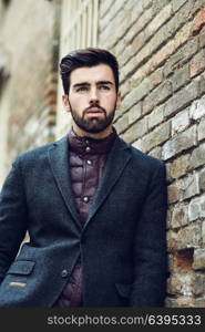Young bearded man, model of fashion, standing in urban background wearing british elegant suit. Guy with beard and modern hairstyle in the street.
