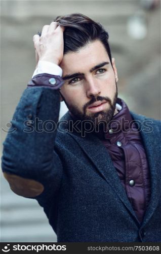 Young bearded man, model of fashion, standing in urban background wearing british elegant suit. Guy with beard and modern hairstyle in the street.. Young bearded man in urban background wearing british elegant suit in the street.