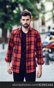 Young bearded man, model of fashion, standing in urban background wearing casual clothes. Guy with beard and modern hairstyle in the street with plaid shirt.