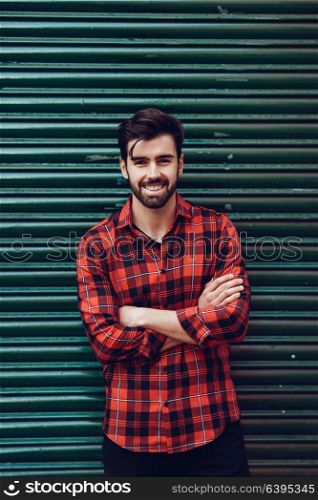 Young bearded man, model of fashion, wearing a plaid shirt with a green blind behind him. Smiling guy with beard and modern hairstyle in urban background.
