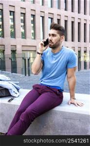 Young bearded man sitting in the street while talking by mobile phone