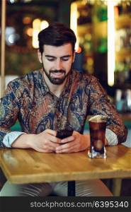 Young bearded man wearing casual clothes looking at his smartphone in a modern pub. Guy with beard and modern hairstyle drinking a cola.