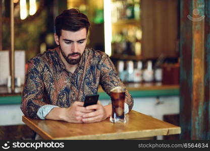 Young bearded man wearing casual clothes looking at his smartphone in a modern pub. Guy with beard and modern hairstyle drinking a cola.