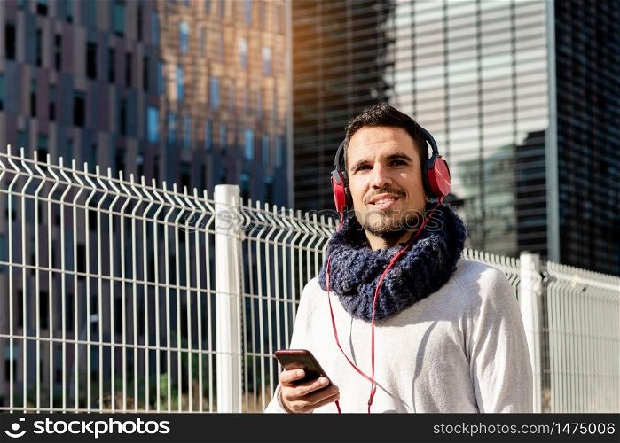 Young bearded man with headphones and holding smartphone while walking against skyscrapers in sunny day