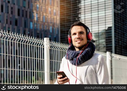 Young bearded man with headphones and holding smartphone while walking against skyscrapers in sunny day