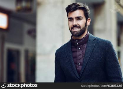 Young bearded smiling man, model of fashion, in urban background wearing british elegant suit. Guy with beard and modern hairstyle in the street.