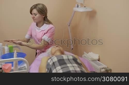 Young beautician moisturizing hands with lotion and massaging senior woman&acute;s face in beauty spa. Relaxed elderly woman receiving face skin therapy in beauty salon. Beauty, spa, cosmetology and relaxation concept.