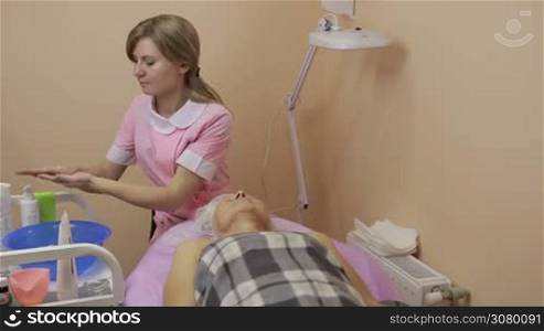 Young beautician moisturizing hands with lotion and massaging senior woman&acute;s face in beauty spa. Relaxed elderly woman receiving face skin therapy in beauty salon. Beauty, spa, cosmetology and relaxation concept.