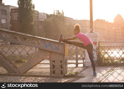 young beautiful African American woman doing warming up and stretching with her leg raised to the bridge before the morning run with the sunrise in the background