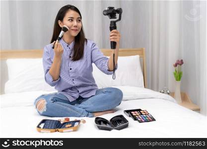 Young beautiful Asian woman beauty vlogger or blogger recording live how to make up tutorial to share on social media using Gimbal Stabilizer on mobile phone. Using for vlog social media influencer concept.