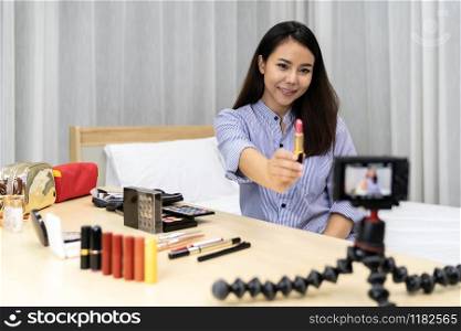 Young beautiful Asian woman beauty vlogger or blogger recording live how to make up tutorial to share on social media using digital camera on table tripod. Using for vlog social media influencer concept.