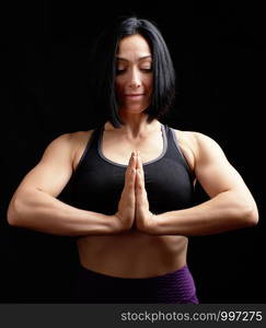 young beautiful athletic girl in a black top, hands makes a gesture of namaste with joined hands in front of the chest, black background