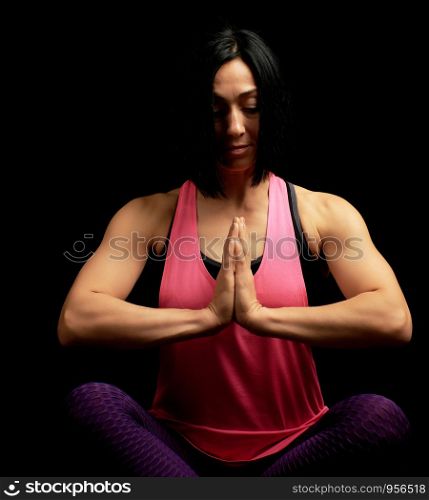 young beautiful athletic girl in a pink top sits with crossed legs in a lotus position, hands makes a gesture of namaste with joined hands in front of the chest, black background