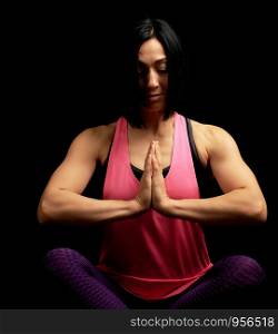 young beautiful athletic girl in a pink top sits with crossed legs in a lotus position, hands makes a gesture of namaste with joined hands in front of the chest, black background