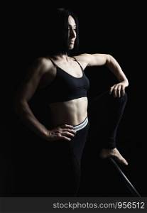 young beautiful athletic girl with muscles in black uniform posing sideways, leg raised up, studio photo in low key
