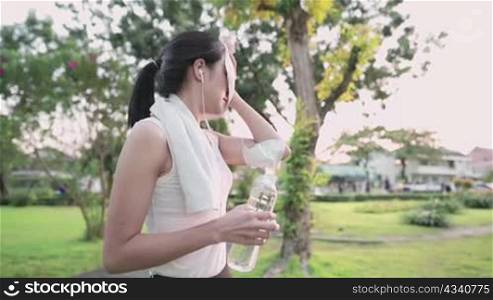 Young beautiful athletic woman directly drinks mineral from reusable bottle with wipes sweat using white towel from her face and neck after jogging in green beautiful park, feeling fresh and relax
