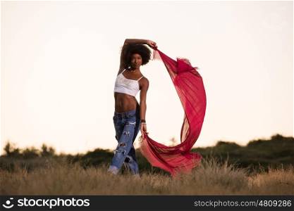 Young beautiful black girl laughs and dances outdoors with a scarf in her hands in a meadow during sunset