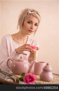 Young beautiful blond woman eating macaroon while having tea-party. She is very satisfacted. Short hair and pink colors - modern style