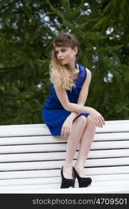Young beautiful blonde girl in blue dress sitting on a bench in summer park