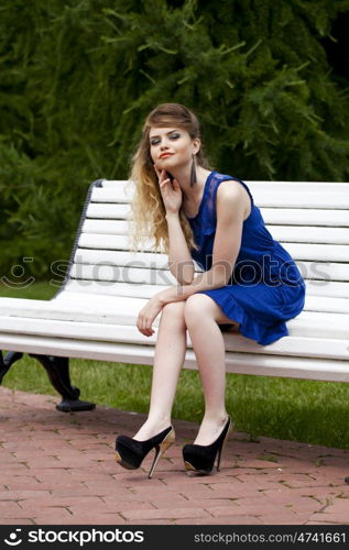 Young beautiful blonde girl in blue dress sitting on a bench in summer park