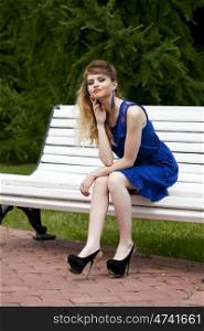 Young beautiful blonde girl in blue dress sitting on a bench in summer park