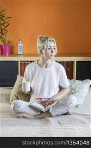 Young beautiful blonde-haired female is sitting on bed with a cup of coffee wearing pajamas in domestic atmosphere