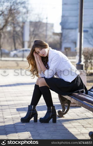 Young beautiful blonde schoolgirl sitting on a bench, spring street