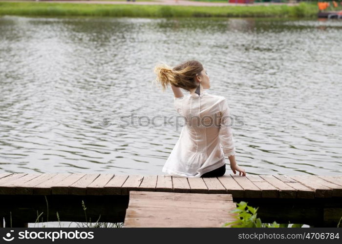 Young beautiful blonde woman in a white tunic sits on a wooden pier on the lake