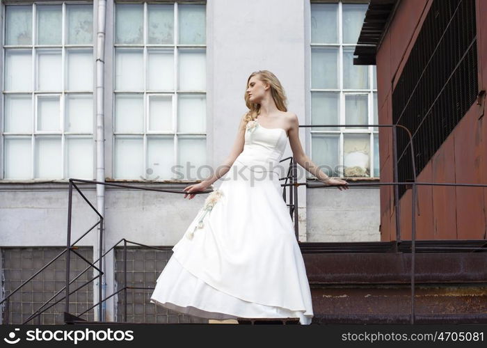 Young beautiful blonde woman in bridal dress posing on the old iron stairs to the roof of a tall building