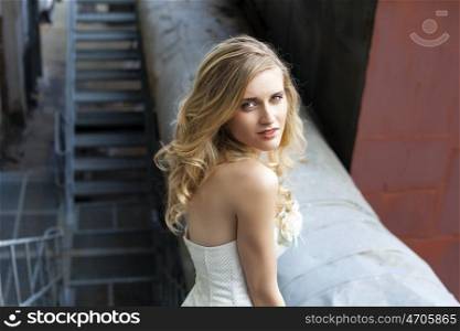 Young beautiful blonde woman in bridal dress posing on the old iron stairs to the roof of a tall building