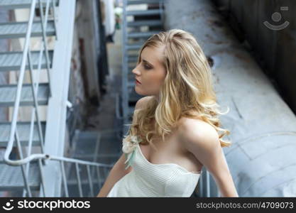 Young beautiful blonde woman in bridal dress posing on the old iron stairs to the roof of a tall building