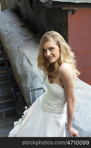 Young beautiful blonde woman in bridal dress posing on the old iron stairs to the roof of a tall building