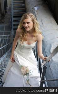 Young beautiful blonde woman in bridal dress posing on the old iron stairs to the roof of a tall building