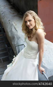 Young beautiful blonde woman in bridal dress posing on the old iron stairs to the roof of a tall building