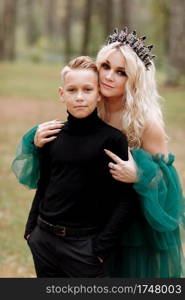 young beautiful blonde woman queen with young boy in black outfit. Princess mother walks with son. autumn green forest mystic. Vintage medieval shiny crown. Long evening green dress. magic fantasy.. young beautiful blonde woman queen with young boy in black outfit. Princess mother walks with son. autumn green forest mystic. Vintage medieval shiny crown. Long evening green dress. magic fantasy