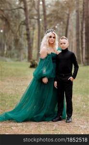 young beautiful blonde woman queen with young boy in black outfit. Princess mother walks with son. autumn green forest mystic. Vintage medieval shiny crown. Long evening green dress. magic fantasy.. young beautiful blonde woman queen with young boy in black outfit. Princess mother walks with son. autumn green forest mystic. Vintage medieval shiny crown. Long evening green dress. magic fantasy