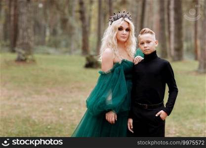young beautiful blonde woman queen with young boy in black outfit. Princess mother walks with son. autumn green forest mystic. Vintage medieval shiny crown. Long evening green dress. magic fantasy.. young beautiful blonde woman queen with young boy in black outfit. Princess mother walks with son. autumn green forest mystic. Vintage medieval shiny crown. Long evening green dress. magic fantasy