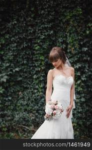 Young beautiful bride in an elegant dress is standing on the field near the forest and holding bouquet of pink flowers and greens with ribbon at nature. Outdoors. After the wedding ceremony.