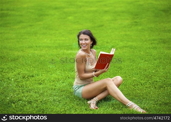 Young beautiful brunette girl with book lying on the green grass in the summer park